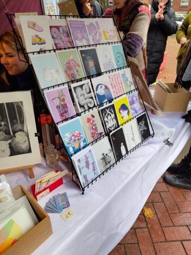 Ein Postkartenständer voll mit verschiedenen bunten Postkarten. Auf einem Marktstand mit noch anderen Kunstdrucken im Bild